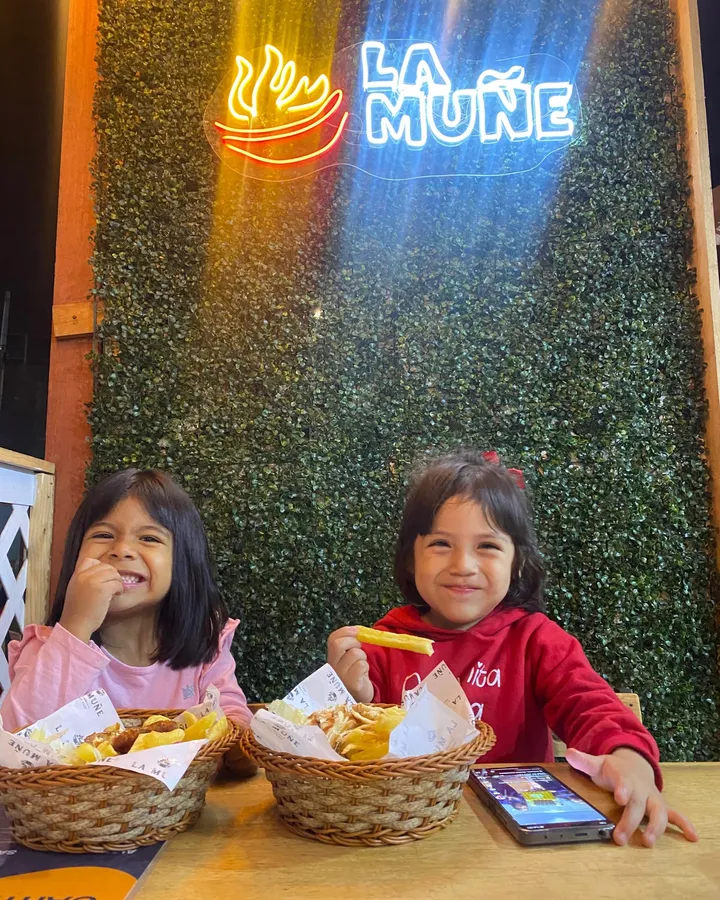 Dos niñas comiendo papitas bajo el letrero de neón de La Muñe